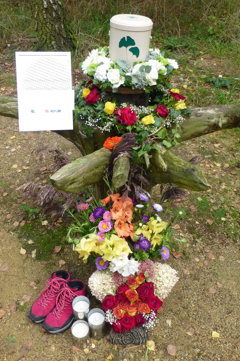 Eine weiße urne auf einem Baumstumpf im Wald umgeben von Blumen, drei Kerzen und einem Paar Laufschuhen Eine weiße urne auf einem Baumstumpf im Wald umgeben von Blumen, drei Kerzen und einem Paar Laufschuhen