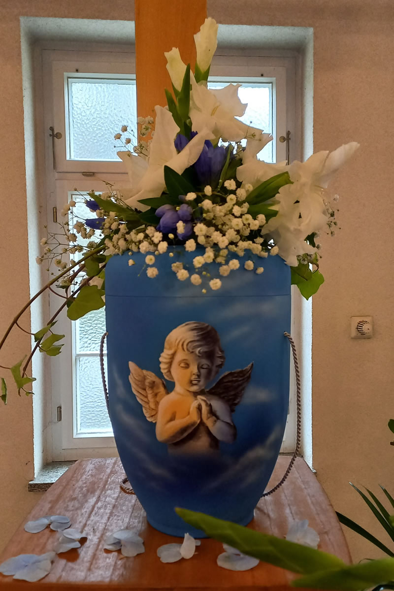 Eine hellblaue Urne mit einem Engel auf der Vorderseite stehend auf einem Tisch vor einem Fenster Eine hellblaue Urne mit einem Engel auf der Vorderseite stehend auf einem Tisch vor einem Fenster